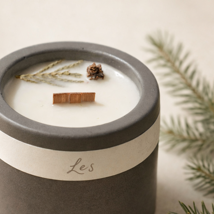 Ručně vyráběná sojová svíčka s vůní lesa v matné keramické nádobě, s dřevěným knotem a přírodními detaily, focená na světlém krémovém pozadí s jemně rozostřenou lesní větvičkou. vyfoceno v detailu