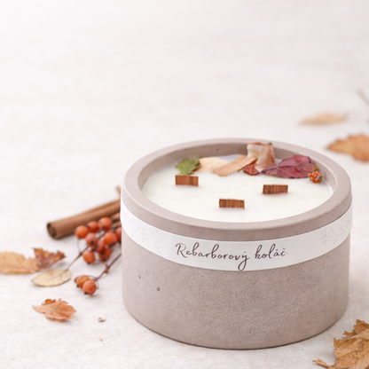 Sójová svíčka Rebarborový koláč ve starorůžovém betonovém obalu s třemi dřevěnými knoty, aranžovaná s podzimním listím, skořicí a sušenými jeřabinami na světlém minimalistickém pozadí.
