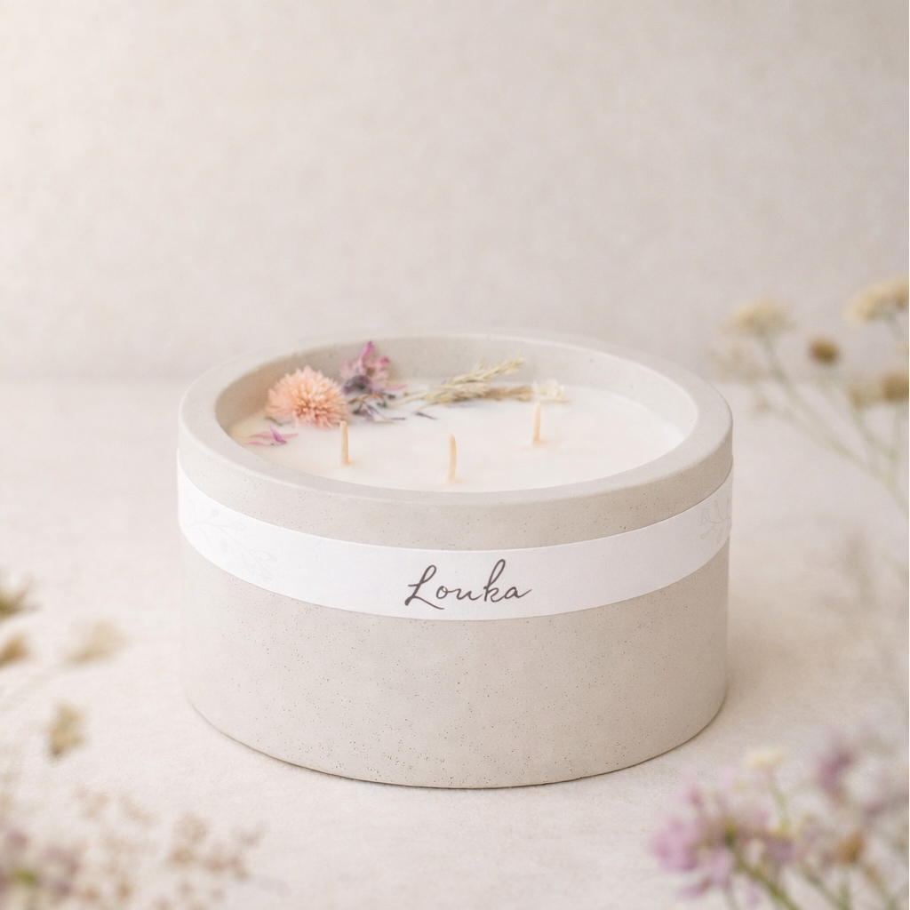 Sójová svíčka Louka v matném světle šedém betonovém obalu, umístěná bokem na světlém povrchu, obklopená jemnými lučními květinami v popředí i pozadí, v měkkém přirozeném světle s hřejivým, minimalistickým a přírodním vzhledem.
