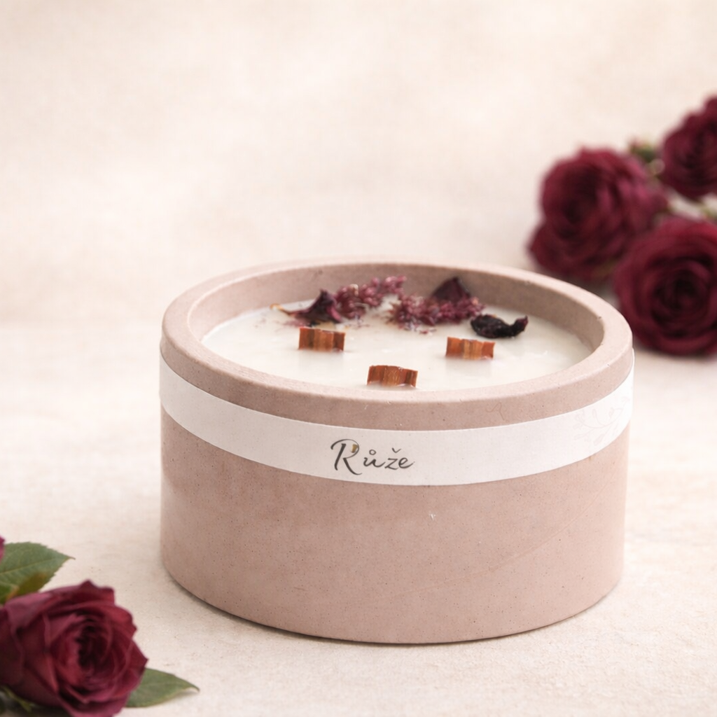 Minimalistická sójová svíčka s vůní růže v matném starorůžovém betonovém obalu, se třemi dřevěnými knoty, aranžovaná s vínově červenými růžemi na světlém krémovém pozadí v jemném přirozeném světle.
