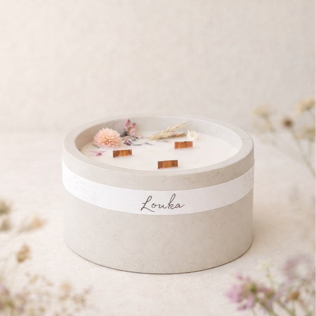 Sójová svíčka Louka v matném světle šedém betonovém obalu, umístěná bokem na světlém povrchu, obklopená jemnými lučními květinami v popředí i pozadí, v měkkém přirozeném světle s hřejivým, minimalistickým a přírodním vzhledem.
