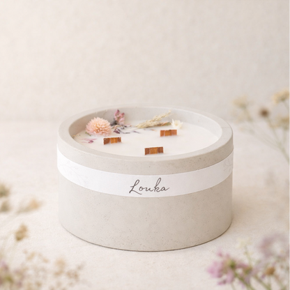 Sójová svíčka Louka v matném světle šedém betonovém obalu, umístěná bokem na světlém povrchu, obklopená jemnými lučními květinami v popředí i pozadí, v měkkém přirozeném světle s hřejivým, minimalistickým a přírodním vzhledem.
