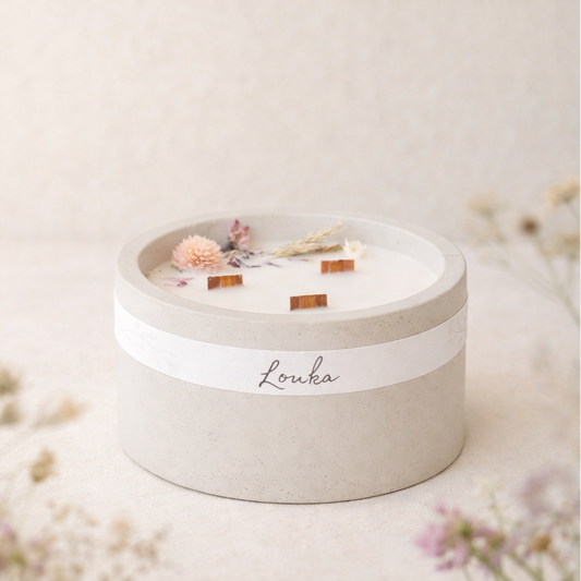 Sójová svíčka Louka v matném světle šedém betonovém obalu, umístěná bokem na světlém povrchu, obklopená jemnými lučními květinami v popředí i pozadí, v měkkém přirozeném světle s hřejivým, minimalistickým a přírodním vzhledem.
