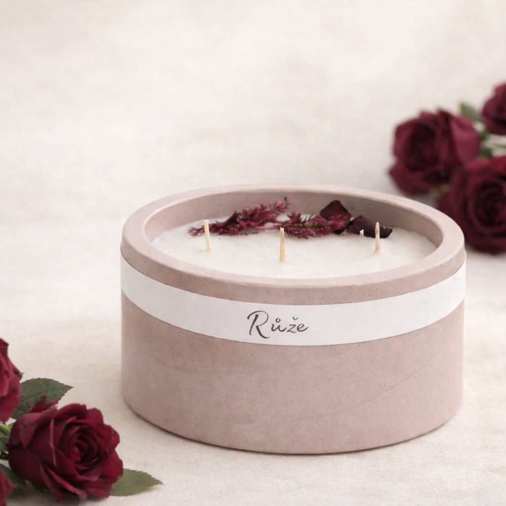 Ručně vyráběná sójová svíčka s vůní růže v matném starorůžovém betonovém obalu se třemi knoty, doplněná vínově zbarvenými růžemi na světlém krémovém pozadí v jemném přirozeném světle.