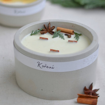 Ručně vyráběná vonná svíčka Koření ve světle  šedé betonové mističce se třemi knoty, ozdobená skořicí, badyánem a větvičkami jehličí.