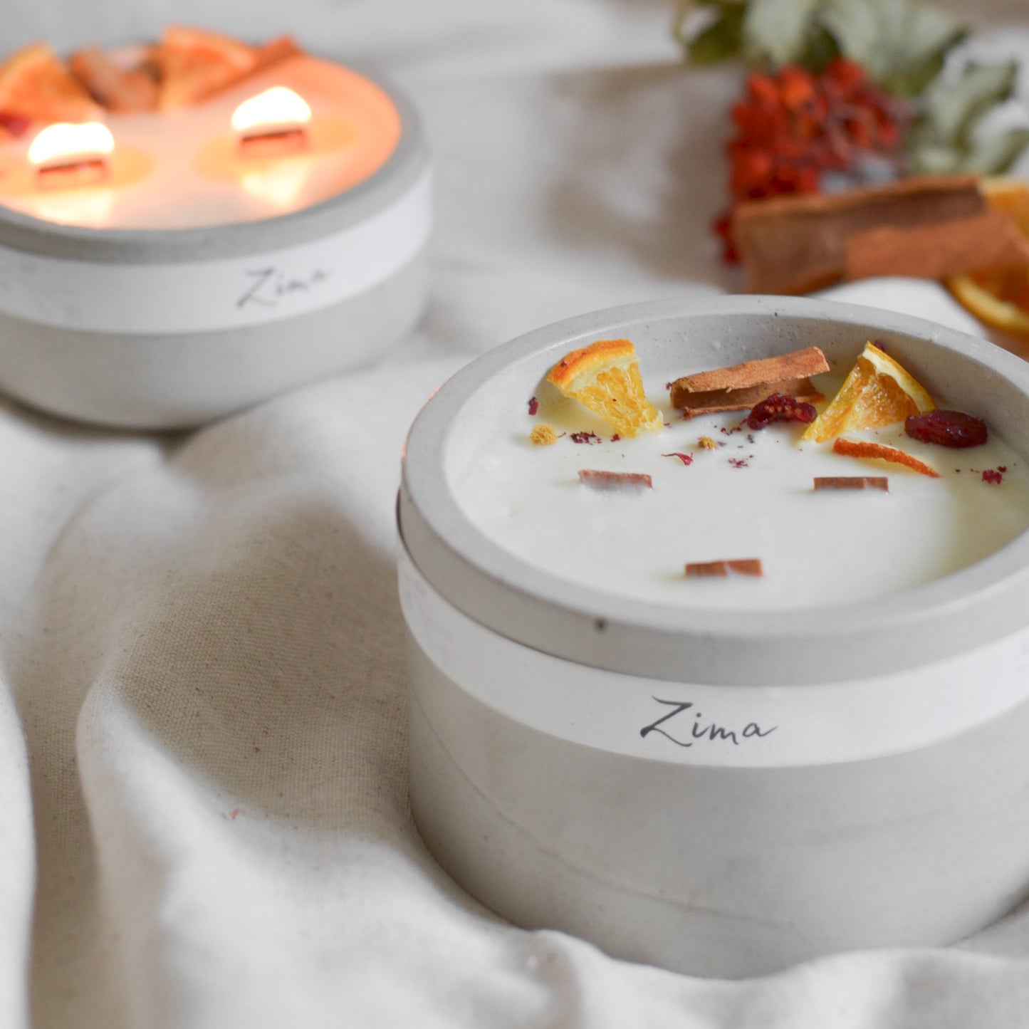 Sójová svicka ve svetle sedem betonu se tremi drvenymi knoty s vuni zimy. Zdobena zimnim korenim a pomerancem. V pozadi svicka hori