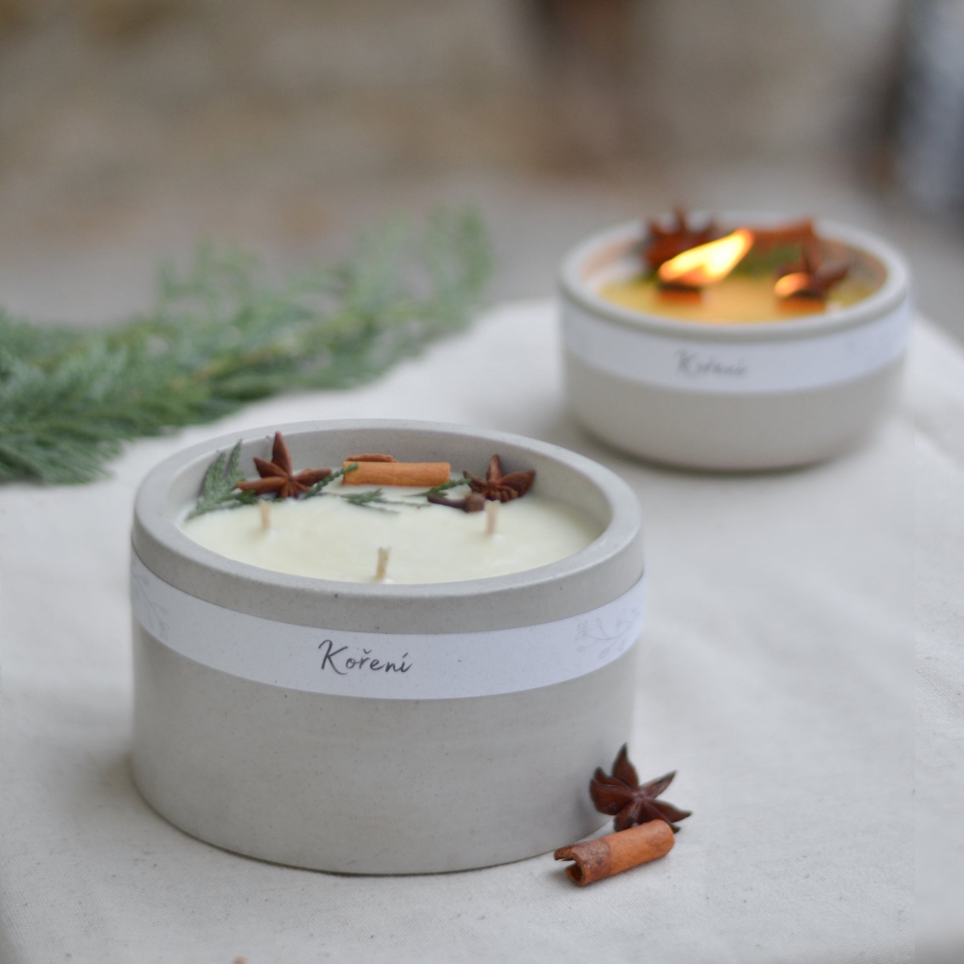 Ručně vyráběná vonná svíčka Koření ve světle  šedé betonové mističce se třemi knoty, ozdobená skořicí, badyánem a větvičkami jehličí.