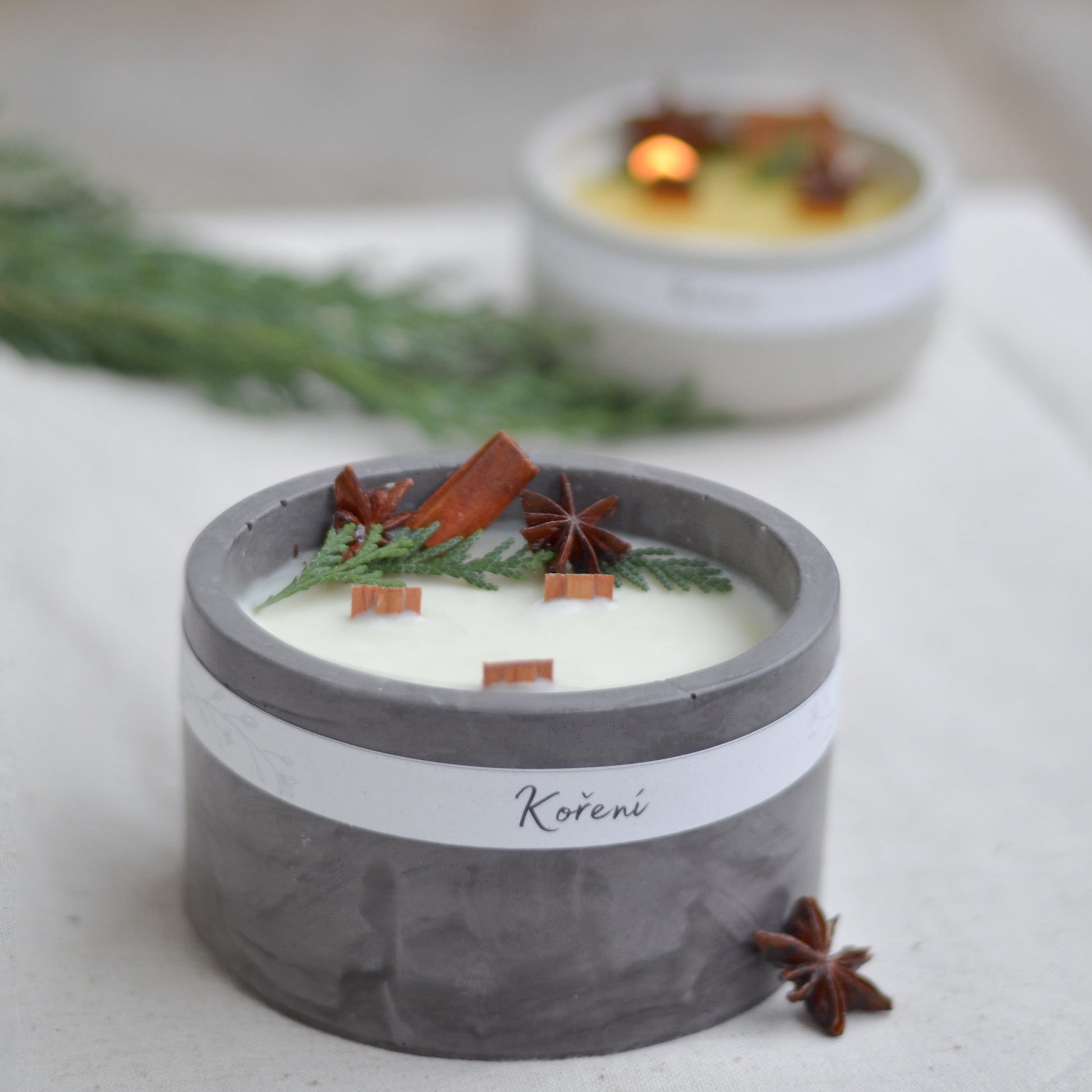 Ručně vyráběná vonná svíčka Koření ve tmavě  šedé betonové mističce se třemi knoty, ozdobená skořicí, badyánem a větvičkami jehličí.