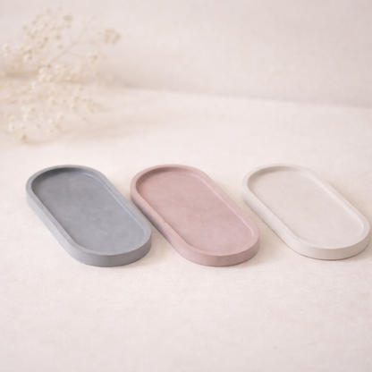 „Tři oválné betonové podtácky v jemných přírodních odstínech šedé, pudrově růžové a krémové, focené na světlém neutrálním pozadí s měkkým přirozeným světlem a jemně rozostřenými sušenými květinami v pozadí.