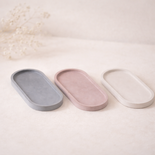„Tři oválné betonové podtácky v jemných přírodních odstínech šedé, pudrově růžové a krémové, focené na světlém neutrálním pozadí s měkkým přirozeným světlem a jemně rozostřenými sušenými květinami v pozadí.