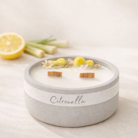 Minimalistická přírodní svíčka Citronella v matné keramické misce s drevenymi  knoty, aranžovaná na světlém pozadí s citronem a stébly citronové trávy, v jemném teplém světle.