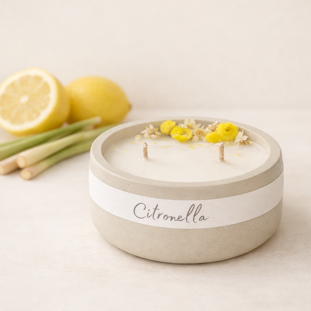 Minimalistická přírodní svíčka Citronella v matné keramické misce s bavlněnými knoty, aranžovaná na světlém pozadí s citronem a stébly citronové trávy, v jemném teplém světle.