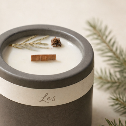 Ručně vyráběná sojová svíčka s vůní lesa v matné keramické nádobě, s dřevěným knotem a přírodními detaily, focená na světlém krémovém pozadí s jemně rozostřenou lesní větvičkou. vyfoceno v detailu