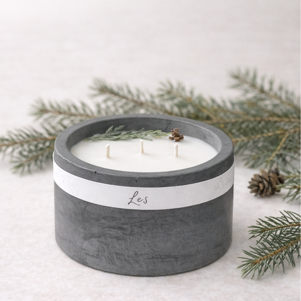 Sójová svíčka Les v tmavě šedém betonovém obalu s třemi bavlněnými knoty, aranžovaná s lesními větvičkami na světlém minimalistickém pozadí.