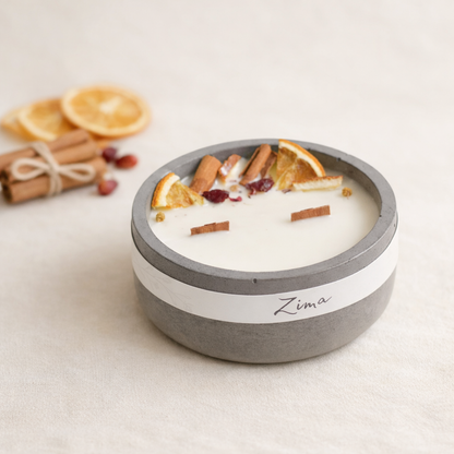 Ručně vyráběná svíčka Zima v matné betonové misce s krémovým voskem, zdobená sušeným pomerančem, skořicí a brusinkami, naaranžovaná v minimalistickém přírodním stylu na světlém béžovém pozadí s měkkým denním světlem.