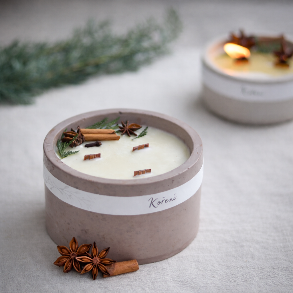 Detailní produktová fotografie ručně vyráběné svíčky v jemně růžovém betonovém obalu s etiketou „Koření“. Povrch svíčky je zdoben skořicí, badyánem a zelenými větvičkami, v popředí leží badyán a kousek skořice. V pozadí je rozostřená zapálená svíčka na světlém textilním podkladu.