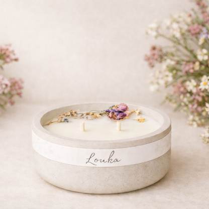 Ručně vyráběná svíčka Louka v matné světlé keramické nádobě se dvěma knoty, zdobená sušenými lučními květy, aranžovaná v jemném přírodním prostředí s rozostřeným pozadím.