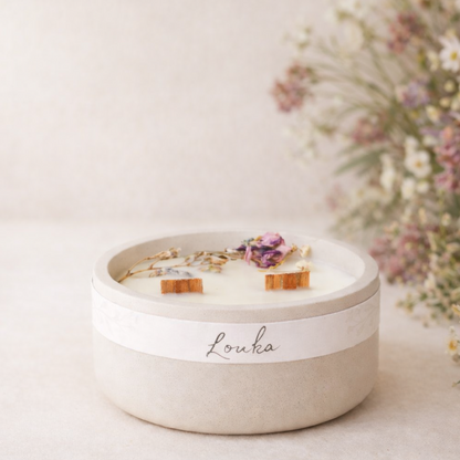 Ručně vyráběná svíčka Louka v matné světlé keramické nádobě se dvěma knoty, zdobená sušenými lučními květy, aranžovaná v jemném přírodním prostředí s rozostřeným pozadím.