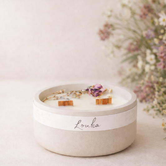 Ručně vyráběná svíčka Louka v matné světlé keramické nádobě se dvěma knoty, zdobená sušenými lučními květy, aranžovaná v jemném přírodním prostředí s rozostřeným pozadím.