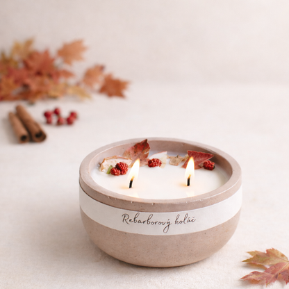 Ručně vyráběná přírodní svíčka Rebarborový koláč v betonove misce s matným povrchem, se dvěma knoty, doplněná sušeným ovocem a podzimním listím v jemně rozostřeném světlém pozadí, s listím soustředěným převážně na levé straně.