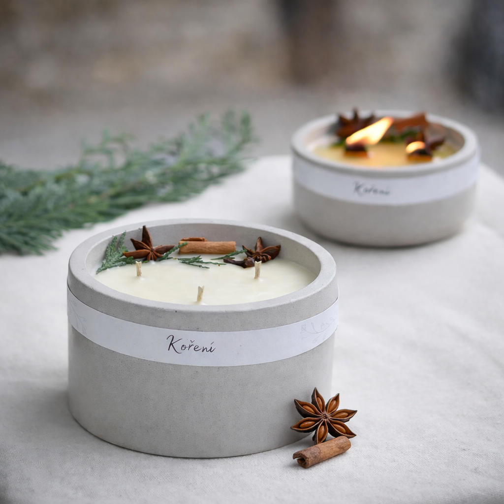 Ručně vyráběná sójová svíčka v šedém betonovém kalíšku s bílou etiketou Koření, zdobená skořicí a jehličím, položená na světlé látce s jemně rozostřeným pozadím.