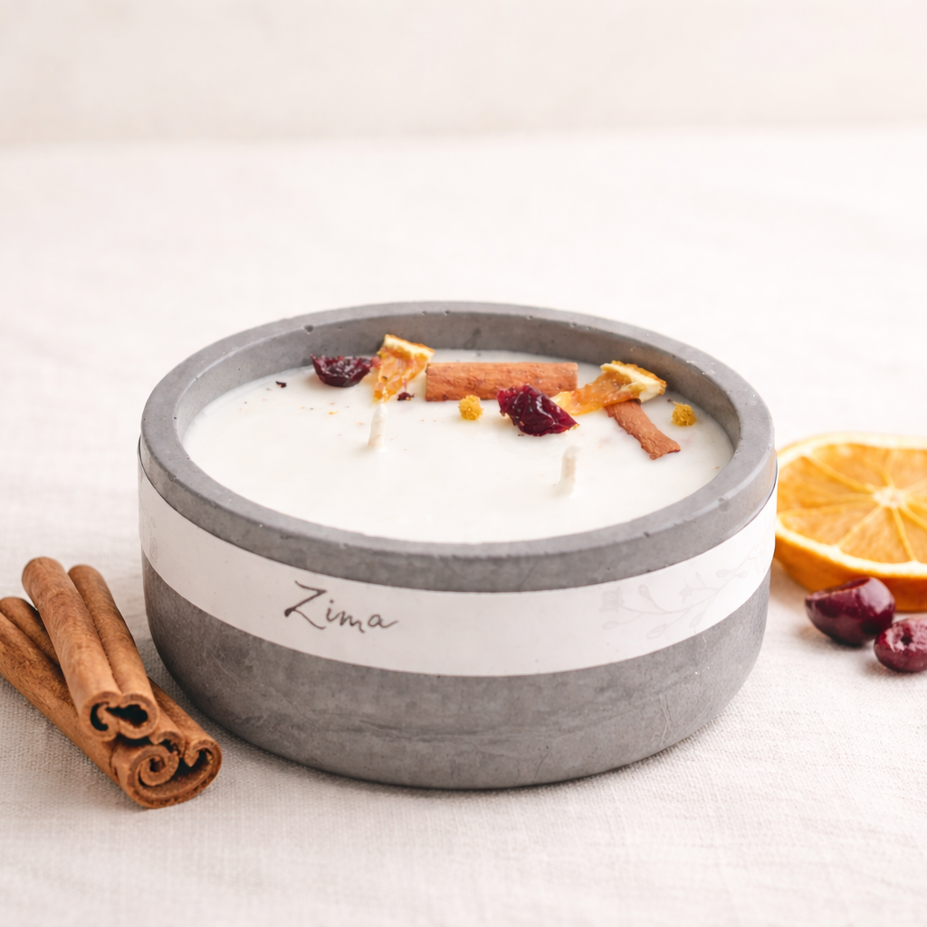 Ručně vyráběná svíčka v šedé betonové misce s bílým voskem, zdobená sušeným pomerančem, brusinkami a skořicí. Svíčka je umístěna na světlém textilním podkladu, v popředí leží skořicové tyčinky a plátek pomeranče. Fotografie má jemné přirozené světlo, teplé tóny a minimalistický přírodní styl.