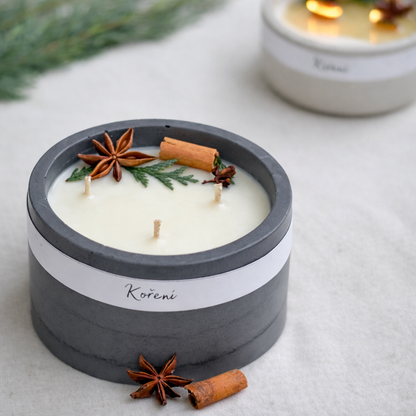 Detailní produktová fotografie ručně vyráběné vonné svíčky v šedém betonovém kalíšku s bílým voskem. Povrch svíčky je zdobený badyánem, skořicí, hřebíčkem a větvičkou jehličnanu. Na obalu je minimalistický štítek s názvem Koření. Svíčka je položena na světlé textilní látce, v pozadí je jemně rozostřená druhá zapálená svíčka a zelené větvičky.