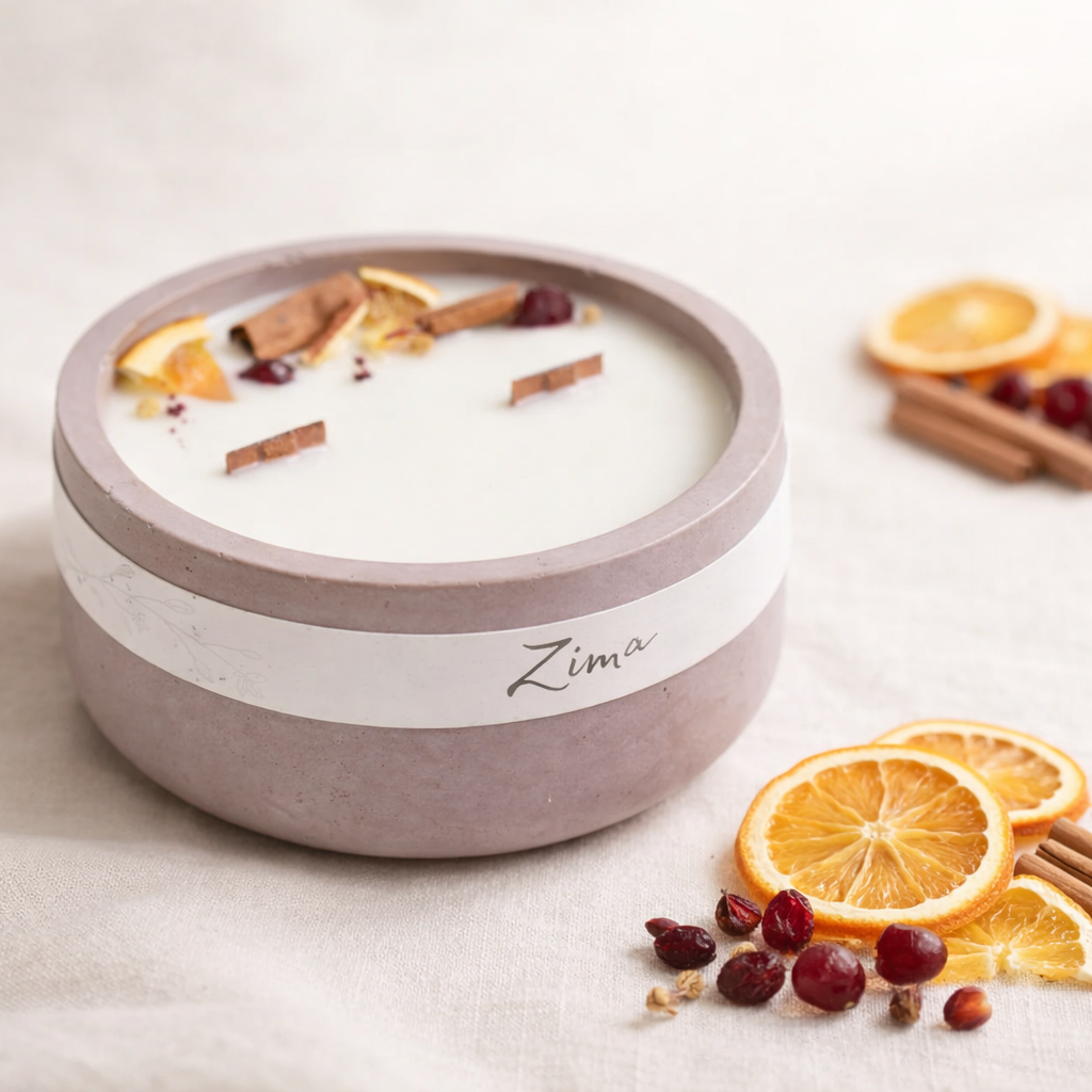 Ručně vyráběná přírodní svíčka v matné betonove misce s jemným štítkem „Zima“, aranžovaná na světlém textilním podkladu. V popředí jsou ostré plátky sušeného pomeranče, brusinky a skořice, zatímco dekorace za svíčkou je jemně rozostřená v teplém, minimalistickém prostředí