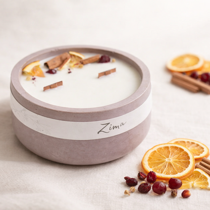 Ručně vyráběná přírodní svíčka v matné betonove misce s jemným štítkem „Zima“, aranžovaná na světlém textilním podkladu. V popředí jsou ostré plátky sušeného pomeranče, brusinky a skořice, zatímco dekorace za svíčkou je jemně rozostřená v teplém, minimalistickém prostředí