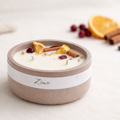 Přírodní ručně vyráběná svíčka Zima v matné keramické nádobě, zdobená sušeným pomerančem, skořicí a brusinkami, nasvícená měkkým teplým světlem na světlém krémovém pozadí.