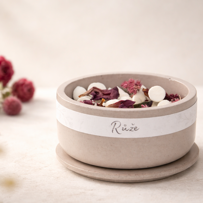 Minimalistická betonová miska v přírodní starorůžové barvě s voskovými kamínky a sušenými vínovými růžemi, nasvícená měkkým přirozeným světlem na světlém krémovém pozadí s rozostřenými růžemi.
