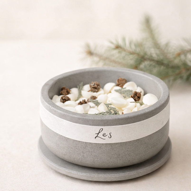 Minimalistická betonová miska s nápisem ‚Les‘ naplněná bílými voskovými kousky, šiškami a lesními bylinami, nasvícená měkkým přirozeným světlem na světlém krémovém pozadí s jemně rozostřenou větvičkou jehličnanu.