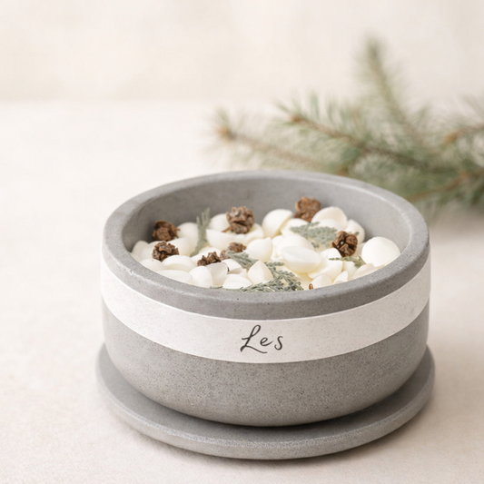 Minimalistická betonová miska s nápisem ‚Les‘ naplněná bílými voskovými kousky, šiškami a lesními bylinami, nasvícená měkkým přirozeným světlem na světlém krémovém pozadí s jemně rozostřenou větvičkou jehličnanu.