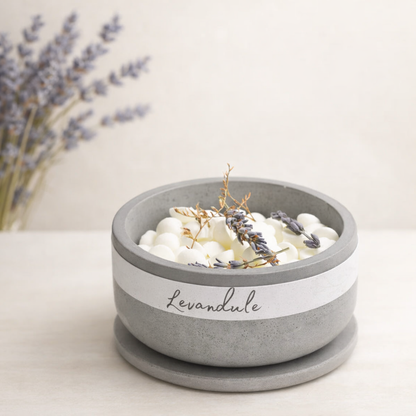 Minimalistická produktová fotografie betonové misky s bílými voskovými peletami a nápisem „Levandule“, doplněné sušenými květy levandule. Produkt je nasvícen měkkým přirozeným světlem, umístěn na světlém povrchu, v pozadí je jemně rozostřená levandule v krémově béžových tónech.