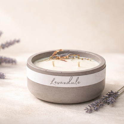 Minimalistická přírodní svíčka Levandule v matné betonové misce, nasvícená měkkým denním světlem, s jemně rozostřeným krémovým pozadím a levandulí aranžovanou z jedné strany.