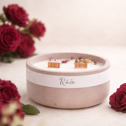 Sójová svíčka s vůní růže v starorůžovém betonovém kalíšku, ruční výroba, doplněná tmavě vínovými růžemi na světlém přírodním pozadí.