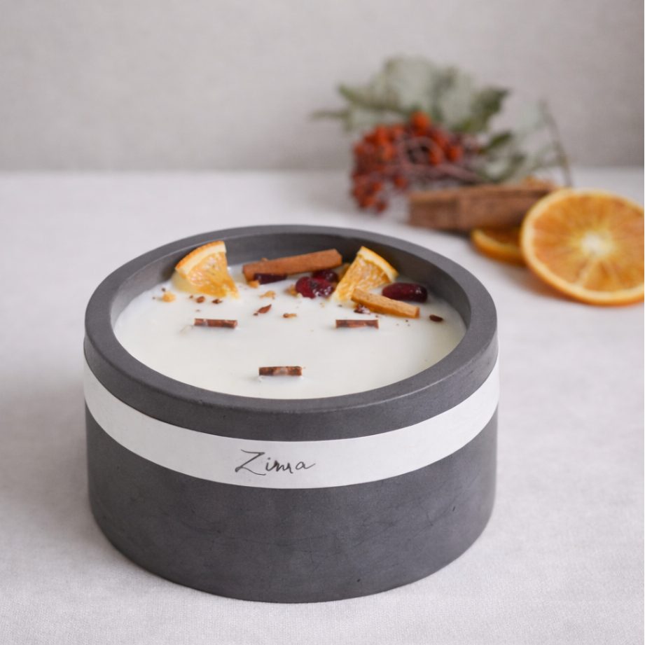 Tmavě šedá betonová svíčka Zima s bílým voskem, zdobená sušenými plátky pomeranče, skořicí a brusinkami, položená na světlé textilii; v pozadí hoří druhá svíčka a zimní dekorace.