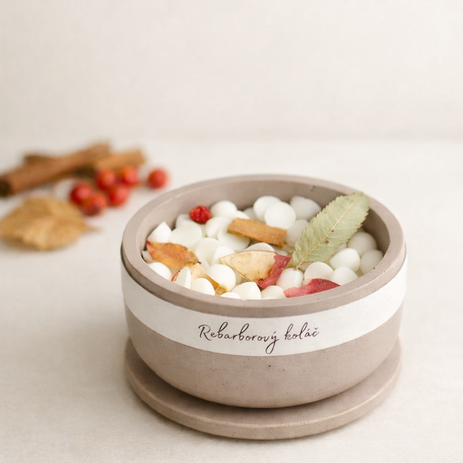 Minimalistická produktová fotografie ručně vyráběné keramické misky s vonnými vosky, s nápisem „Rebarborový koláč“, umístěné na světlém béžovém pozadí. Miska je naplněna bílými voskovými kousky a přírodními dekoracemi z listí, skořice a červených bobulí, s jemným teplým světlem a rozostřeným přírodním pozadím evokujícím podzimní atmosféru.